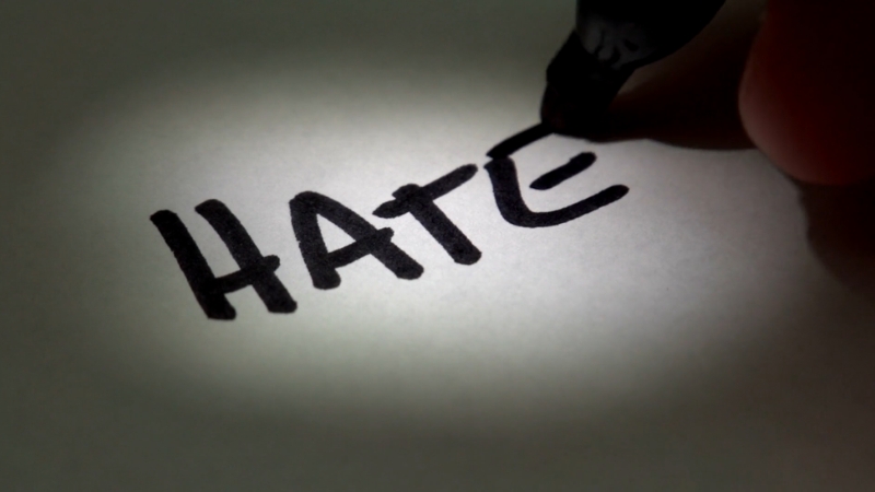 A hand writing the word "HATE" with a marker, illuminated by a spotlight, representing "words that people hate"