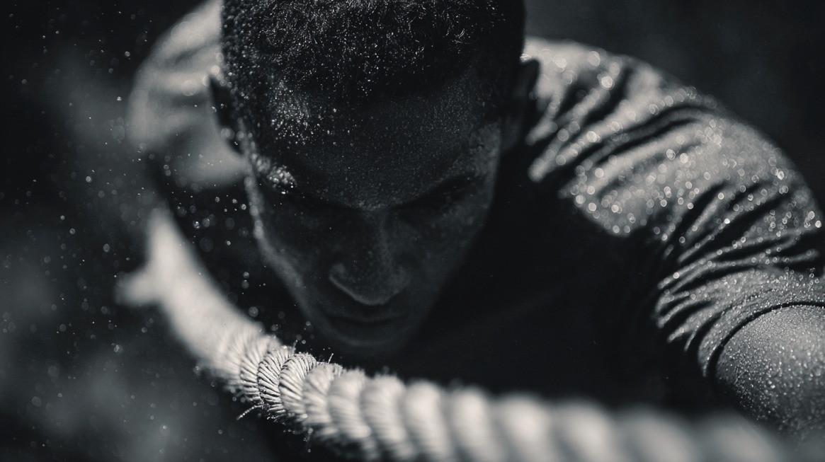 Close-up black and white image of a person covered in sweat, pulling a thick rope with intense focus and determination