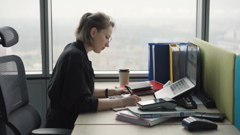 A woman sits at an office desk working with documents and a laptop beside a window